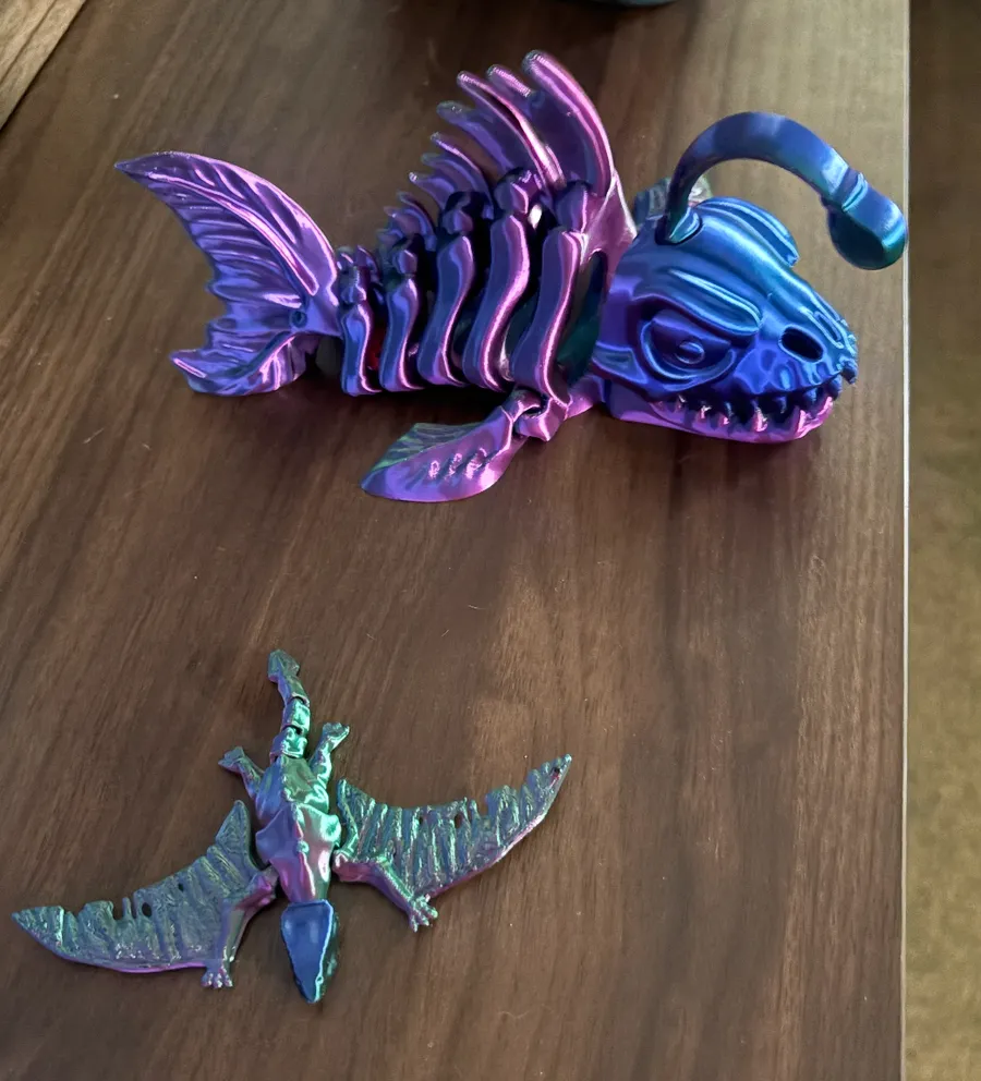 Articulated skeleton angler fish and pterodactyl 3D prints on a wooden table, printed in color-shift filament that fades between purple, blue, and green