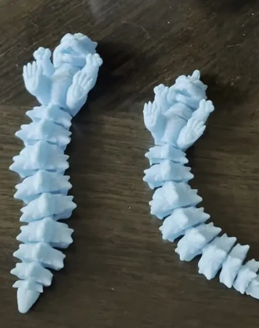 Two blue articulated spike dragon 3D prints lying on a wooden table next to the printer
